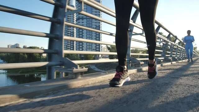 Two sunlit sportswomen jogging along city bridge across river during healthy leisure activity. Fit female runners enjoying cardio training at nature surrounded by high-rise buildings