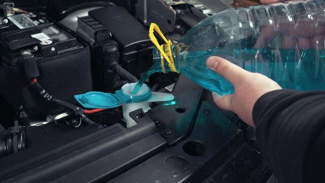 Pouring blue liquid windshield guard from plastic bottle into car, close up. Washer fluid for washing car glass.
