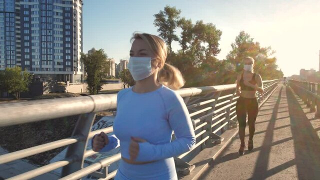 Masked urban joggers going in for sports during coronavirus epidemic. Active sportswomen in protective face masks and tight outfit running along sidewalk of city bridge amid covid-19 pandemic