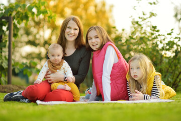 Fototapeta premium Mother and three children having fun on autumn day in city park. Adorable baby boy being held by his mommy. Two older sisters hugging their mom and baby brother. Big family.