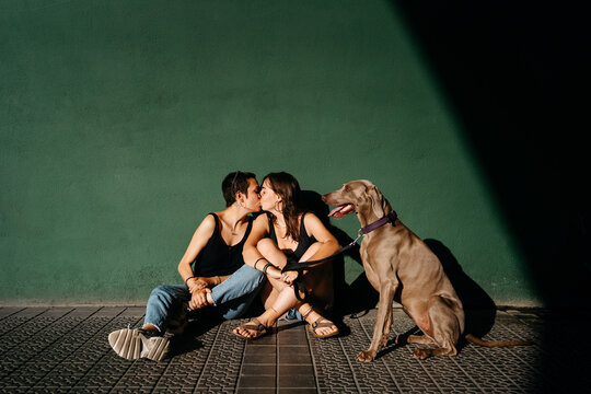 Young lesbian couple with dog kissing near green wall