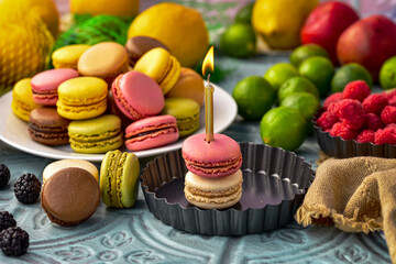tabletop still life with fruit, plate of assorted fruit flavor macaron cookies, and two macarons stacked with gold birthday candle lit with flame