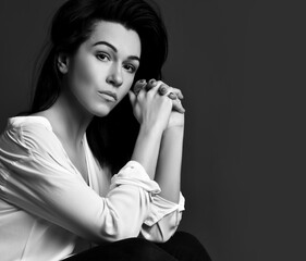 Portrait of Beautiful serious brunette woman in stylish white shirt and trousers sitting looking at camera over dark background. Fashion, stylish casual look for lady concept