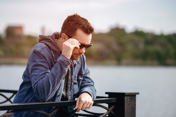 A man in sunglasses stands on a bridge by the river. Stylish guy dressed in denim clothes. Backlight and free space for text