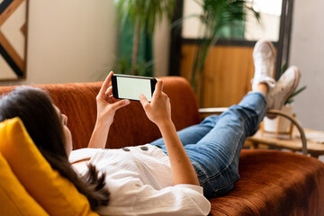Young woman at home lying down in the couch using smartphone .