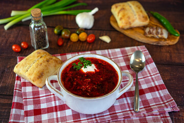 Borscht with sour cream in a white plate on a wooden table. The table also contains bread, bacon, green onions, peppers and garlic. Traditional Ukrainian and Russian cuisine.