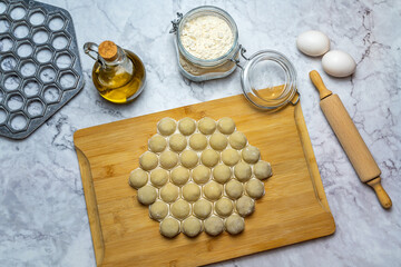 Raw meat dumplings on a cutting board. There are flour, eggs, rolling pin, dumpling maker, olive oil and pepper.