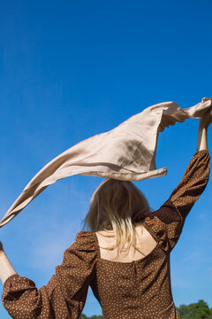 A girl stands with her back against the blue sky in a brown dress and holds a scarf