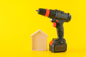 Modern black cordless screwdriver, drill with rechargeable battery and wooden house on yellow...