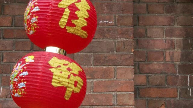Close up shooting of a red chinese lantern decorated with golden ideograms swaying in the wind in font of a red brick building wall.