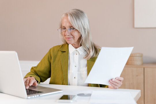 Positive elderly employee reading paper and using laptop
