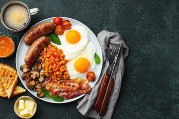 Full English breakfast on a plate with fried eggs, sausages, bacon, beans, toasts and coffee on dark stone background. With copy space. Top view