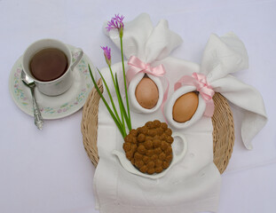 Beautiful festive Easter brunch with eggs in  napkin bunny, cup of tea, cookies and pink flowers on a basket.