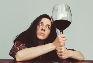 A young girl in a dark plaid shirt dark hair sits at a wooden table and holds a large glass of red wine. Concept of alcohol abuse, alcoholism, hangover, loneliness and depression.