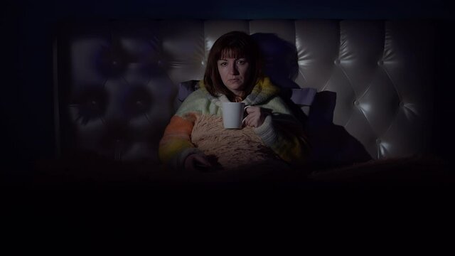 brunette woman on the bed in the evening drinking tea and watching tv with emotions, going through