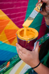 Artist holding paintbrush and paint can with dripping yellow paint next to a geometric colorful wall mural © Leslie Rodriguez