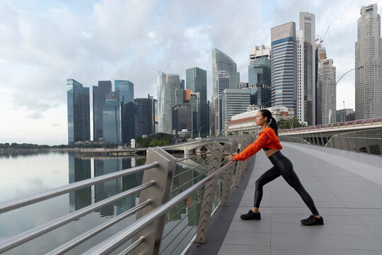 A singaporean woman is stretching on the railing of a bridge. She is wearing an orange hoodie.