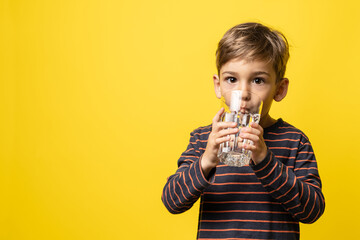 Small caucasian boy little child holding glass of water while standing in front of modern yellow background drinking and looking to the camera with copy space