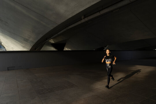 A young asian woman in running outdoors in the morning hours. First lights hits her as she runs under a bridge in the city. Her shirt reads: run.