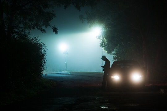 Silhouette of a man, leaning on a car, next to street lights.