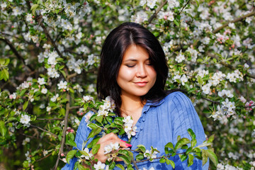 A young attractive plus-size woman with dark hair in a blue dress stands with her eyes closed in a spring blooming garden. Spring time.