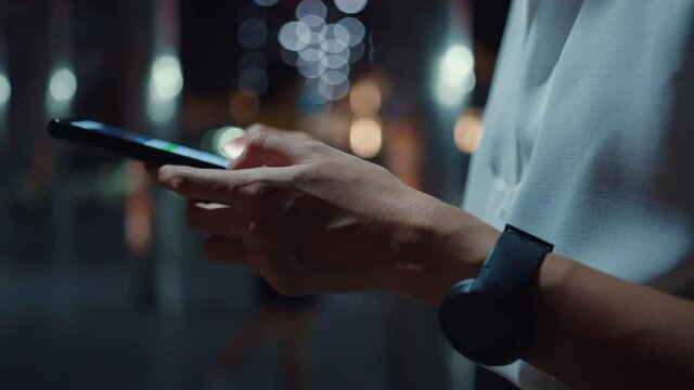 Young Asia businesswoman in fashion office clothes using smart phone typing text message while stand outdoors in urban modern city at night. Business on the go concept. Closeup shot.