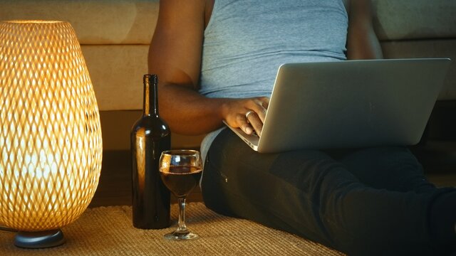 Young African American man using laptop, drinking red wine from glass, looking at the screen. works remotely, work from home concept, freelancer.