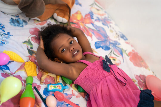 Little Black Girl Lying On The Bed With Toys.