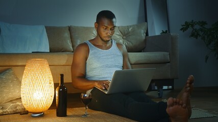 Young African American man using laptop, drinking red wine from glass, looking at the screen. works remotely, work from home concept, freelancer.