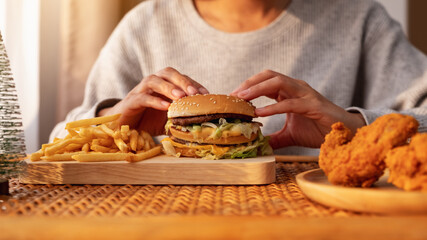 Closeup image of a woman holding and eating hamburger and french fries with fried chicken on the table at home