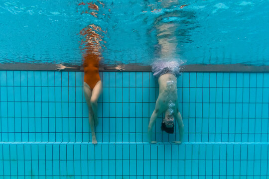 A couple seen underwater in a pool in Singapore. She is standing on the side, he is hanging upside down.