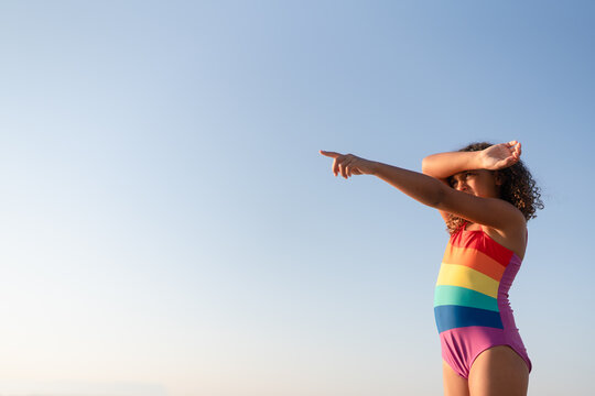 Girl in rainbow swimsuit against blue sky pointing