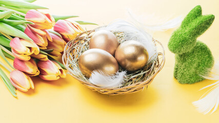 Basket easter decoration: Golden eggs in basket with spring tulips, white feathers on pastel yellow background. Congratulatory easter design.