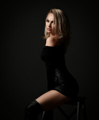 Fototapeta na wymiar Sexy seductive blonde woman vamp in black leather shorts, pullover and boots is sitting sideways looking at camera flirting over dark background. Fashion, style, sexy wear for women concept