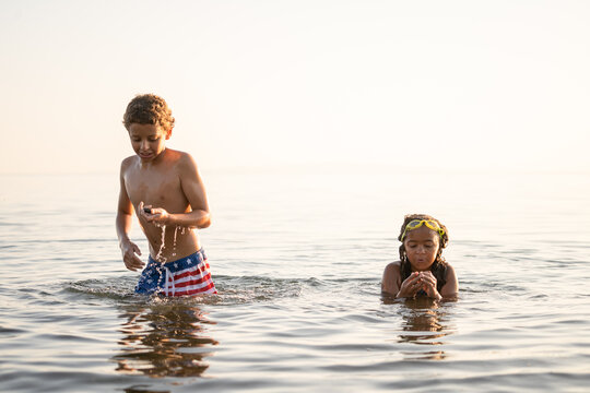 Multiracial Brother And Sister Look At Rocks In The Ocean At Sunset