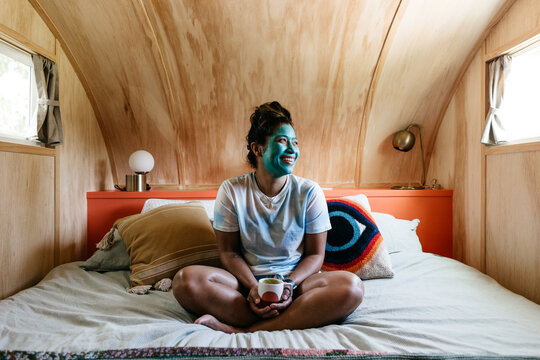 A woman sitting in bed with a cup of coffee and a cleansing face mask on.