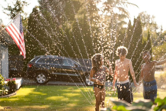 Multiracial siblings play in front yard sprinkler