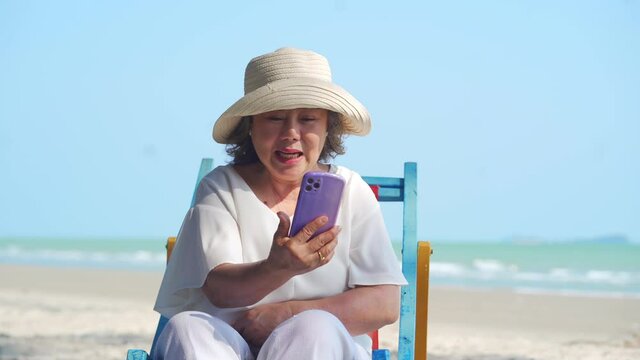 4K Happy Asian senior woman tourist sitting on beach chair by the sea. Healthy elderly retirement female using smartphone for selfie or talking video call with family and friends at tropical beach.