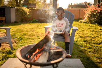 Smiling boy sits in lawn chair looking at campfire