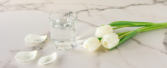 A glass of purified fresh drinking water on a marble table with white tulip flowers . copy space for text