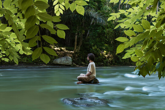 Man practicing meditation while sitting on a small rock in in the Ayung river, on a misty morning in Bali