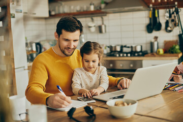 Small girl sitting on father's lap while he is taking notes and working on laptop at home.