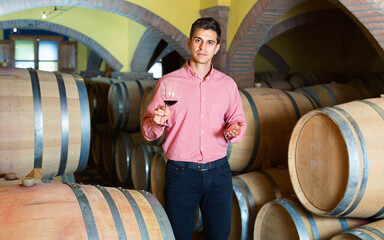 Glad smiling male customer tasting red wine from wooden barrels in factory