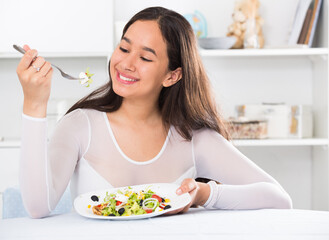 Positive young female eating salad witn fork sitting at home