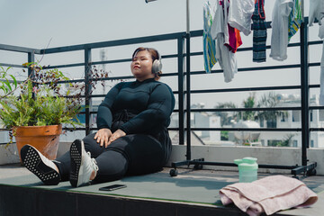 Fototapeta na wymiar Peaceful plus size young asian woman in modern wireless headphones sit relax after doing exercise on mat in balcony, going to loose extra pounds at home, become strong and fit.