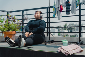 Fototapeta na wymiar Peaceful plus size young asian woman in modern wireless headphones sit relax after doing exercise on mat in balcony, going to loose extra pounds at home, become strong and fit.