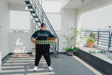 Plus size young asian woman playing hula hoop in balcony. Helps to burn excess fat in body. Strengthens the muscles of the body, going to loose extra pounds at home,