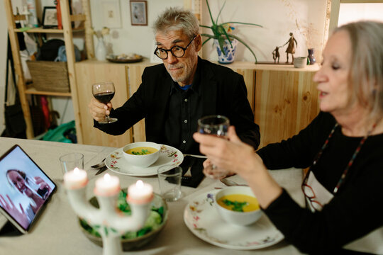 Elderly couple doing a virtual toast with a family member through a tablet set on the table.