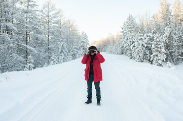 A man in a red jacket stands in a snowy forest on the road in winter and adjusts his snowboard goggles on his face