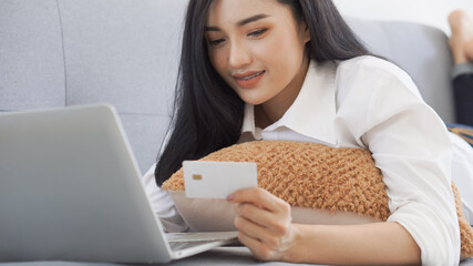 Asian woman holding credit card and using laptop to shopping online, lying on sofa. Shopaholic girl selecting products to cart in online website And pay via internet banking.